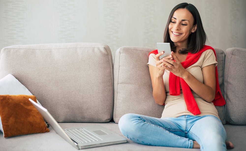 la-mujer-sonriente-confiada-moderna-joven-esta-trabajando-o-relajandose-en-casa-y-sentada-en-el-sofa-mientras-usa-su-telefono-inteligente